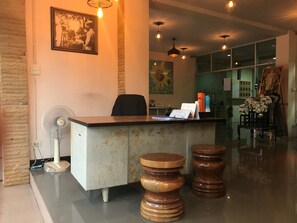 A reception desk with a chair, a fan, and a sign with a phone number. There are wooden stools in front of the desk. - Areechon Apartment (Ban Chang)
