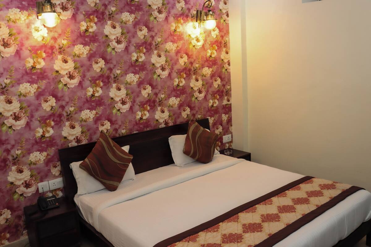 Una cama bien hecha con una colcha blanca y dos almohadones decorativos, una mesita de noche con un teléfono y un fondo de papel tapiz con motivos florales.