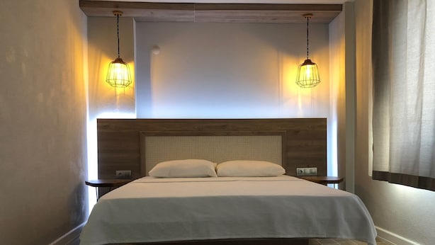 Una habitación de hotel moderna con cabecera de madera, una cama grande con ropa blanca, dos mesitas de noche y dos lámparas colgantes.