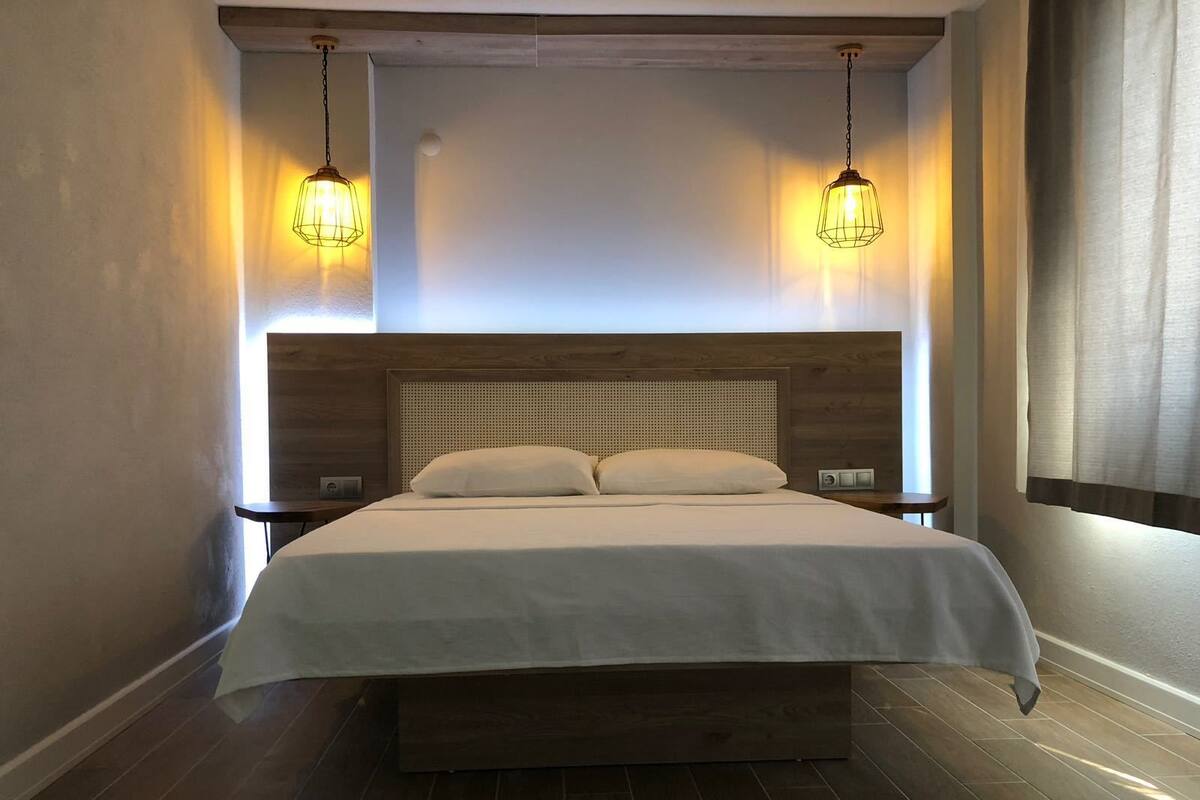 A modern hotel room with a wooden headboard, a large bed with white linens, two bedside tables, and two hanging pendant lights.