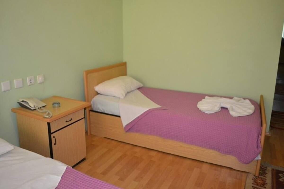 A single-bed room with a wooden headboard, a bedside table with a phone, a small cabinet, and a purple bedspread.