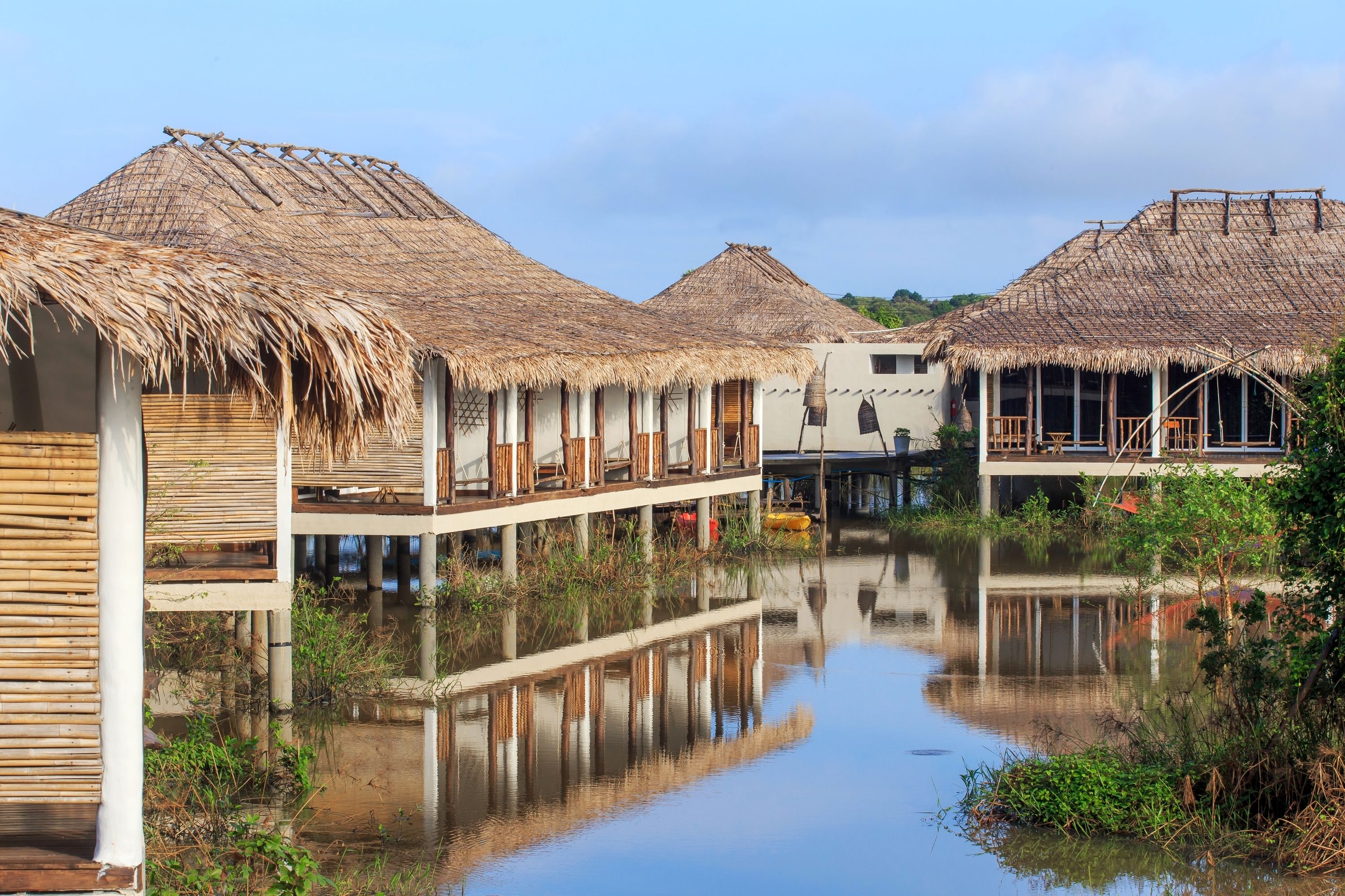 A waterway with thatched-roof buildings, wooden walkways, and a calm reflective surface.