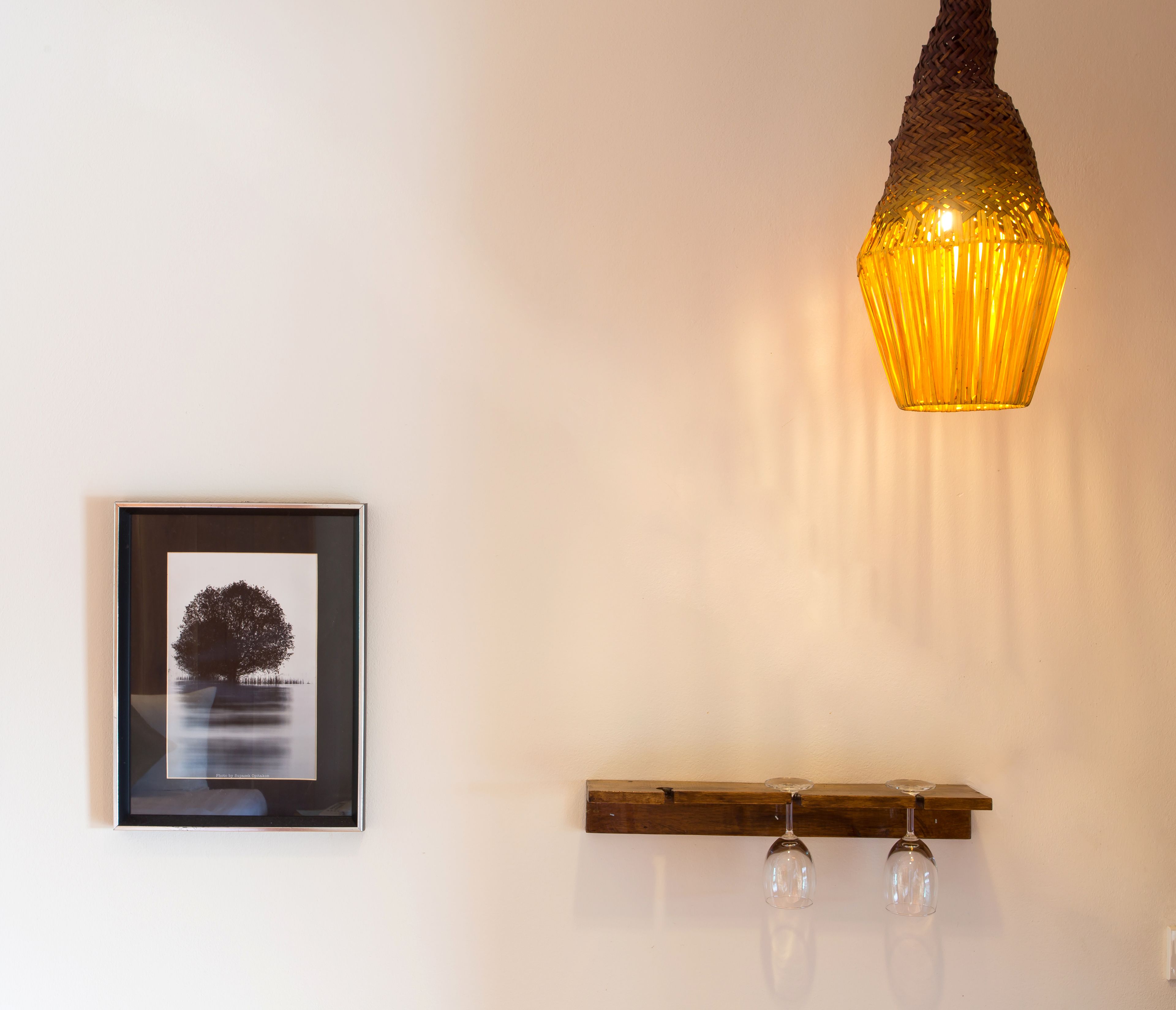 Ein gerahmtes Bild von einem Baum an der Wand, eine Holzablage mit Weingläsern und eine Pendelleuchte.