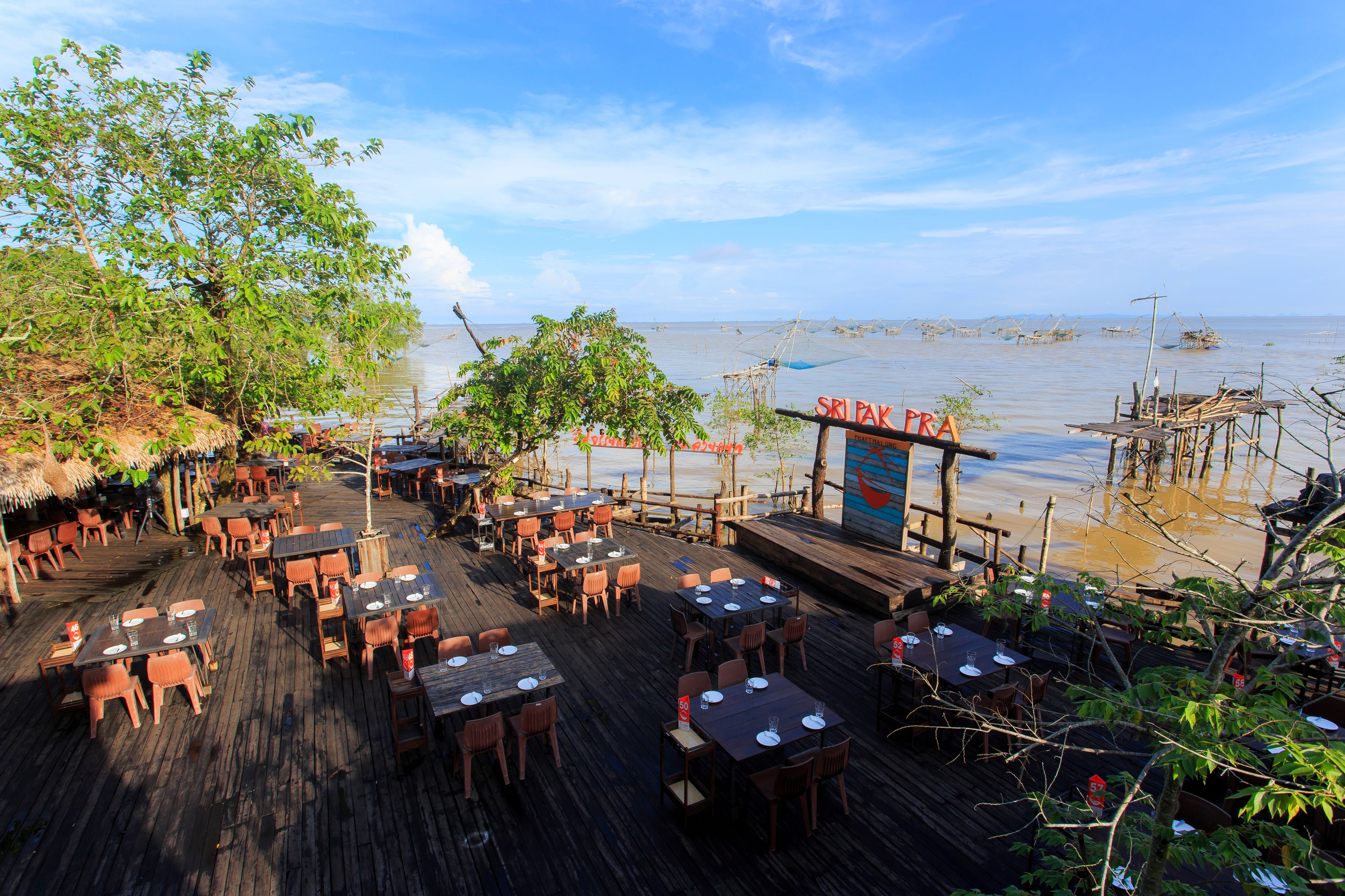 An outdoor restaurant with wooden tables and chairs, a thatched roof, and a view of the sea.