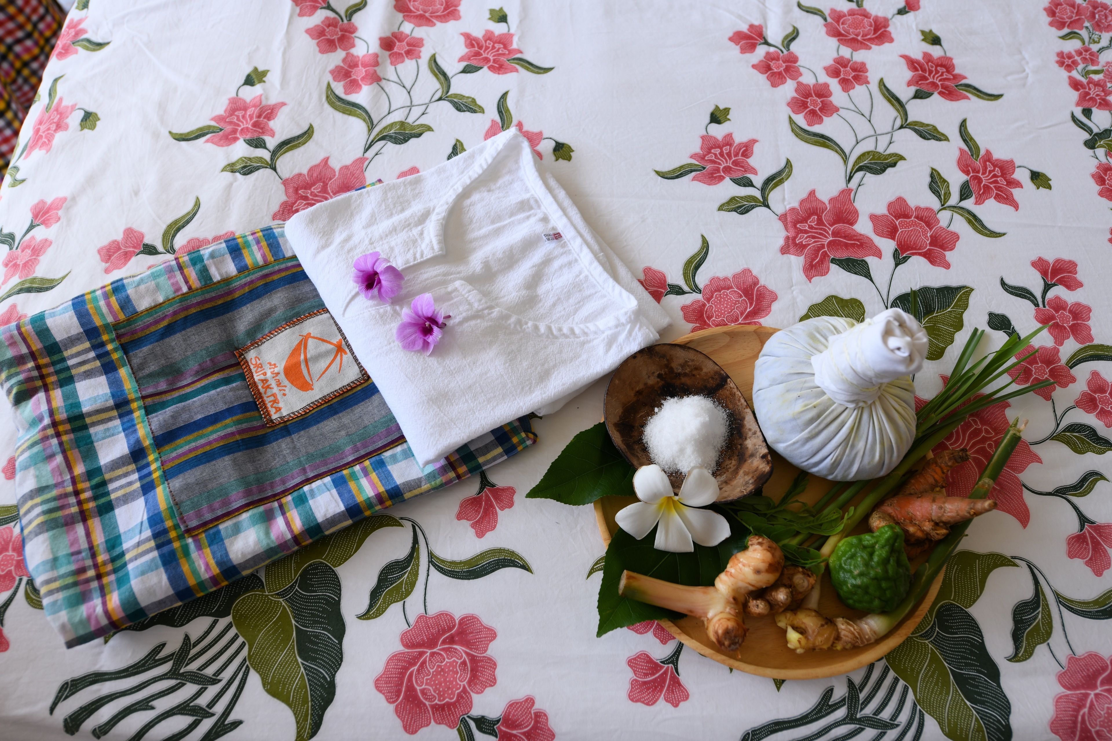 A neatly arranged bed with a floral-patterned bedspread, a folded white towel, a plaid cloth, and a wooden tray with ginger, a white flower, and a small white object.