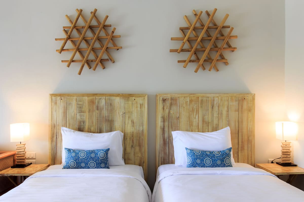 A hotel room with two wooden beds, each with a white mattress and a blue patterned pillow. There are wooden headboards and two wall-mounted wooden decorations.