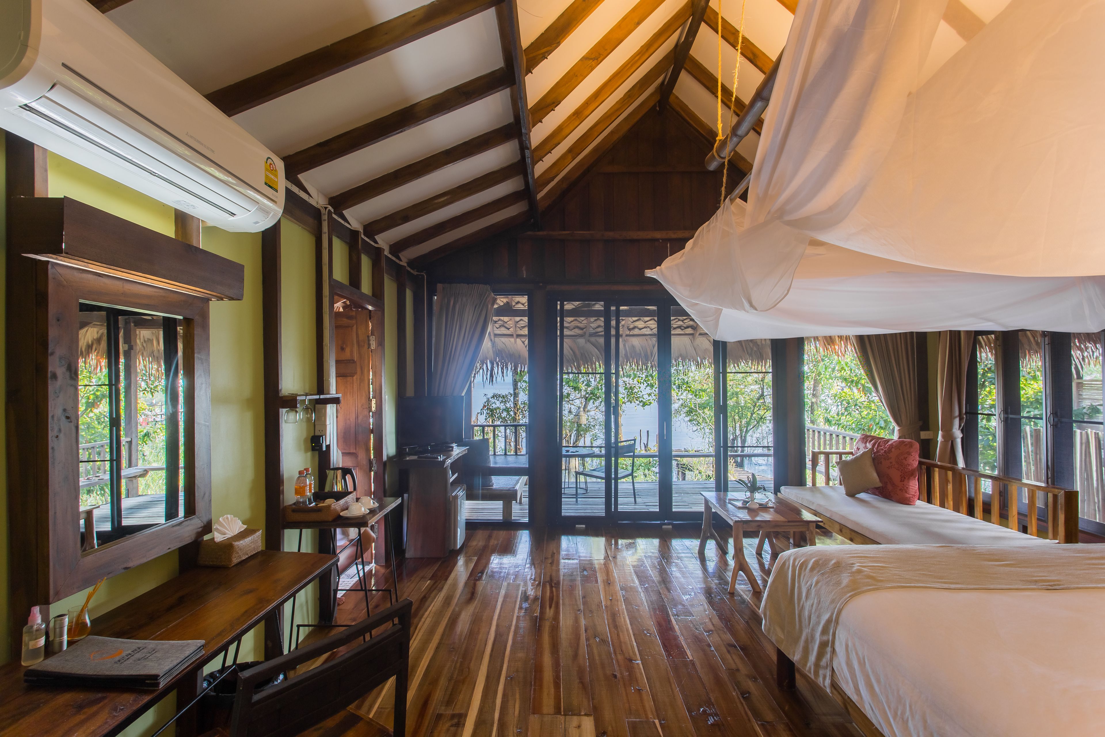 A room with a wooden floor, a bed, a desk with a chair, a window, and a door leading to an outdoor area.