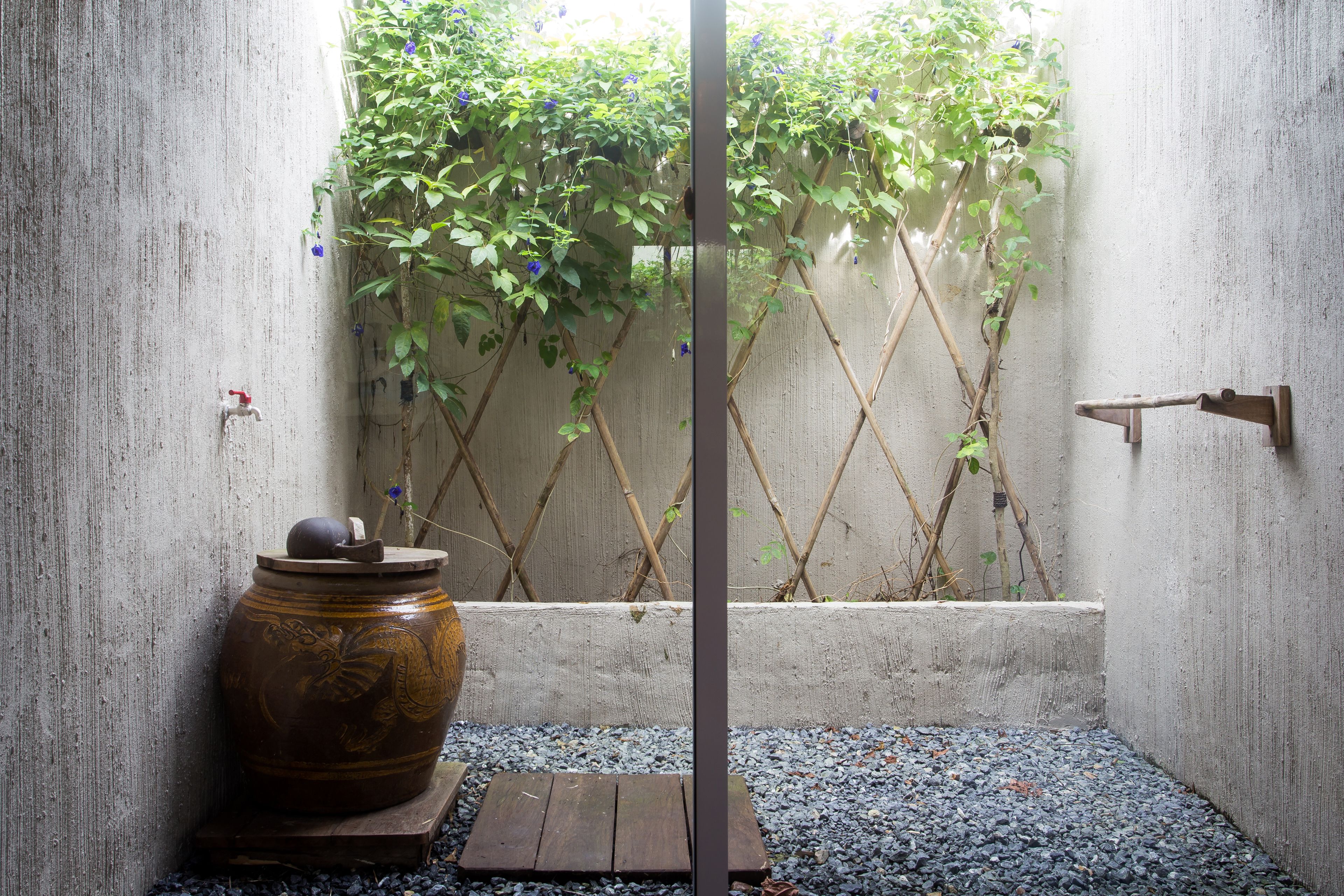 A small, enclosed space with a large ceramic pot, a wooden stool, and a bamboo ladder against a wall with climbing plants.