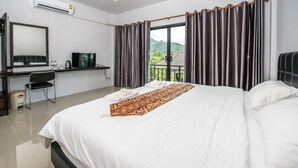 A hotel room with a large bed, a desk with a TV, and a view of greenery outside. - Mr. T Kamala Room (Kamala)