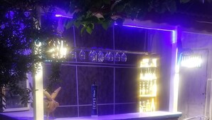 An outdoor bar area with a neon sign, bar stools, and a shelf with bottles.