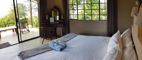 A hotel room with a bed, a desk with a mirror, a chair, and a view of greenery through large windows.