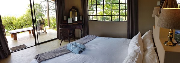 Habitación de hotel con una cama, un escritorio con espejo, una silla y vistas a zonas verdes a través de amplias ventanas.