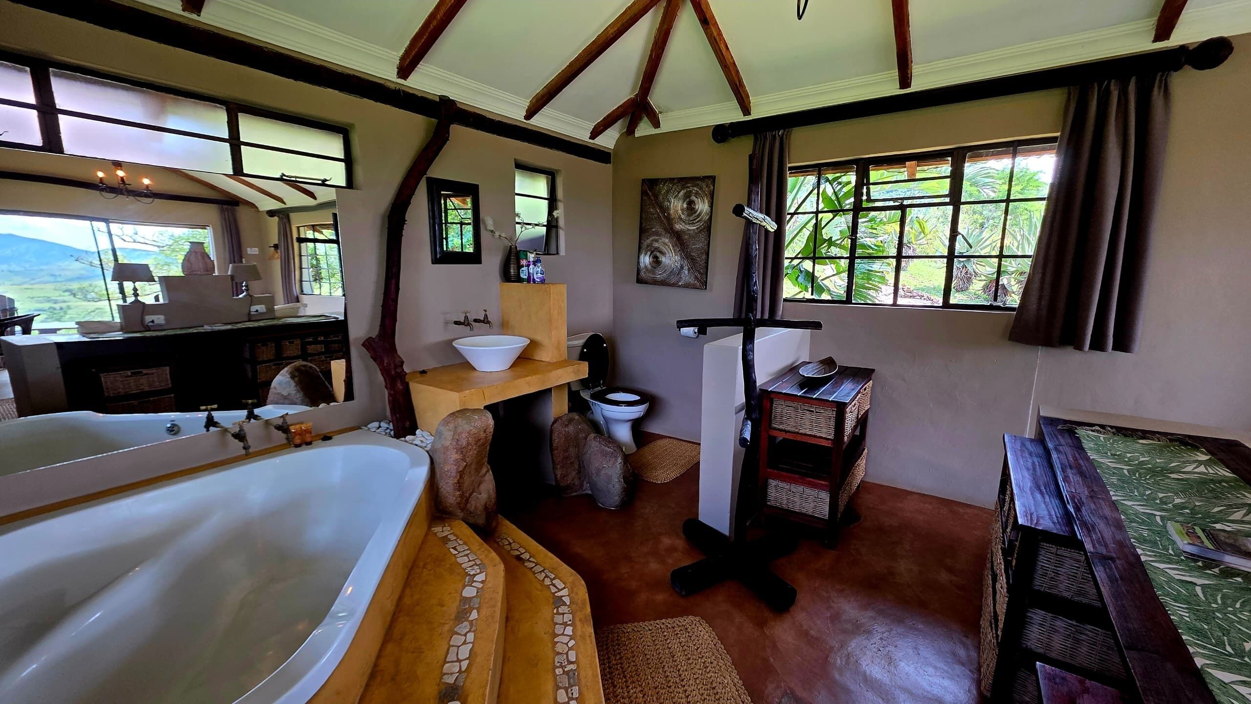 A spacious bedroom with a large bed, a wooden vanity, a sink, and a window with a view of greenery.