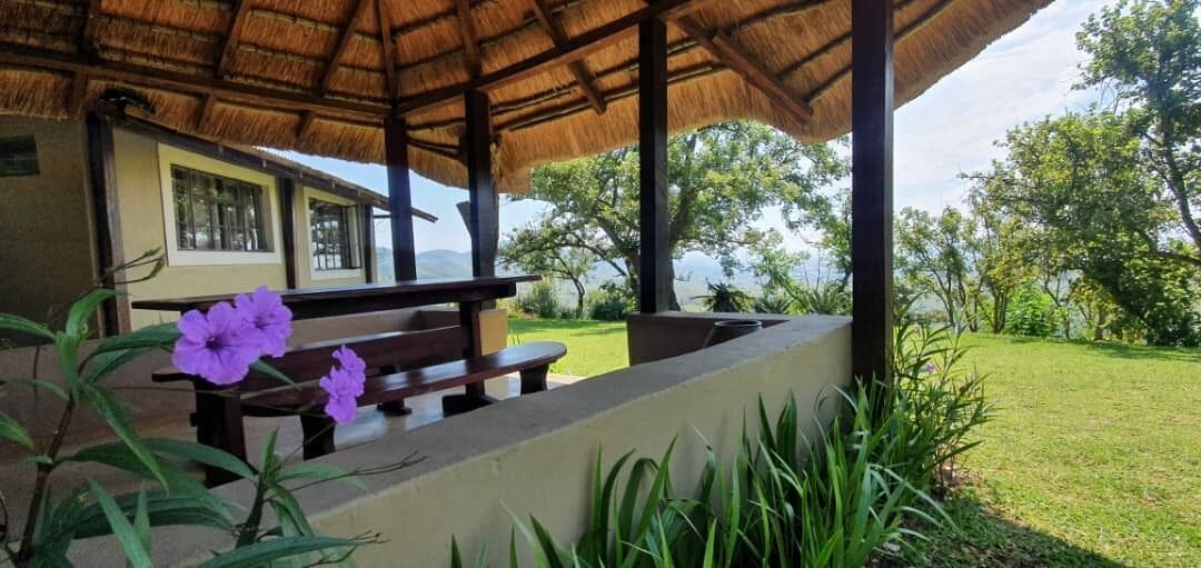 Un espacio al aire libre con techo, bancos y una mesa, rodeado de vegetación y vistas a una casa.