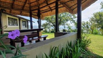 Un espacio al aire libre con techo, bancos y una mesa, rodeado de vegetación y vistas a una casa.