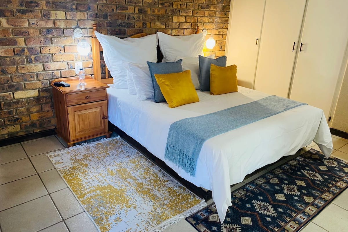 A bedroom with a brick wall, a wooden nightstand, a bed with white bedding and multiple pillows, a blue throw blanket, and a patterned rug.