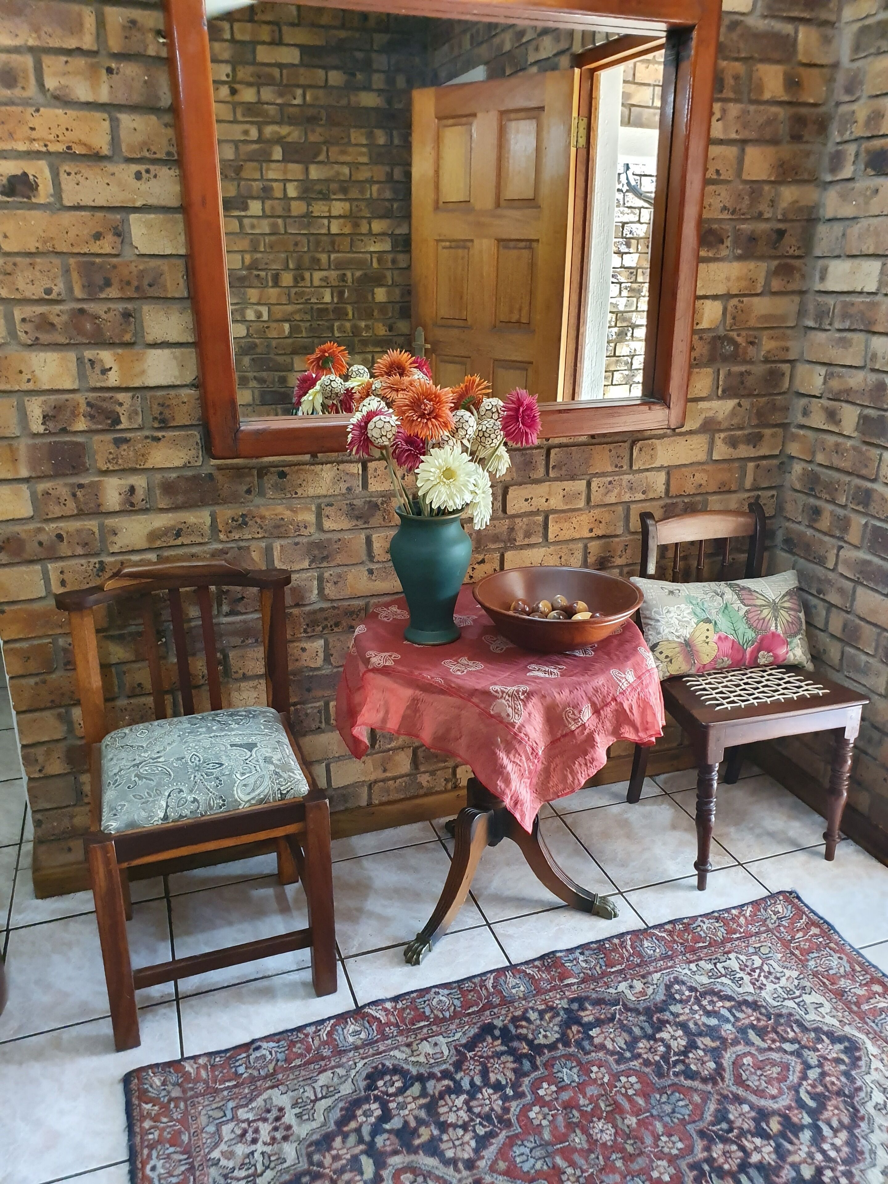 A cozy room with a brick wall, a wooden door, a mirror, a table with a vase of flowers, two chairs, and a patterned rug.