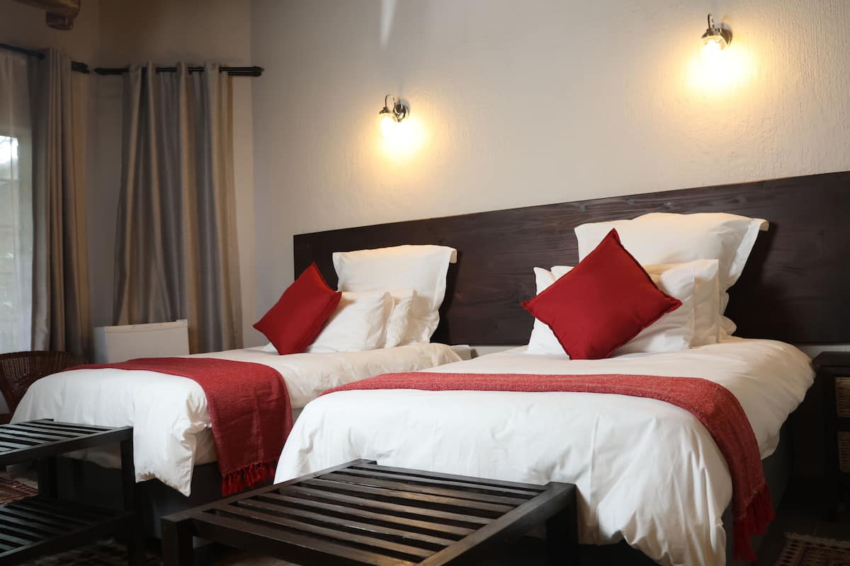 A hotel room with two beds, each with white bedding and red pillows, a dark headboard, and a small table in front of each bed.
