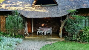 Uma casa com telhado de palha e fachada de tijolo, uma mesa central com cadeiras e um caminho pavimentado que leva à entrada.