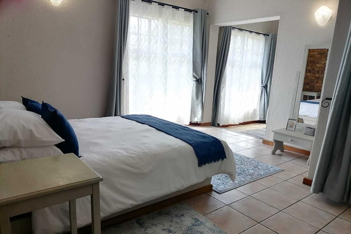 A bedroom with a bed, a bedside table, a mirror, and a view of the outside through large windows.