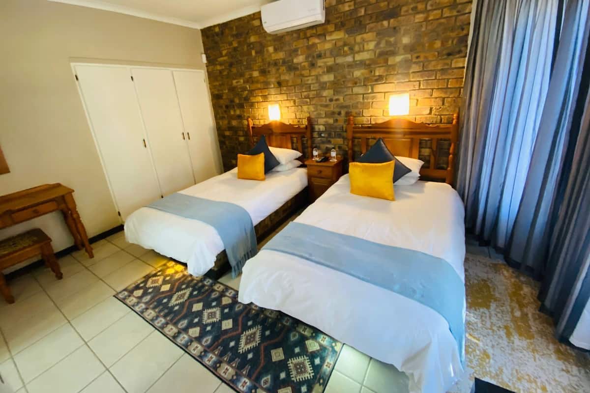 A hotel room with two beds, a wooden desk, a patterned rug, and a brick wall.