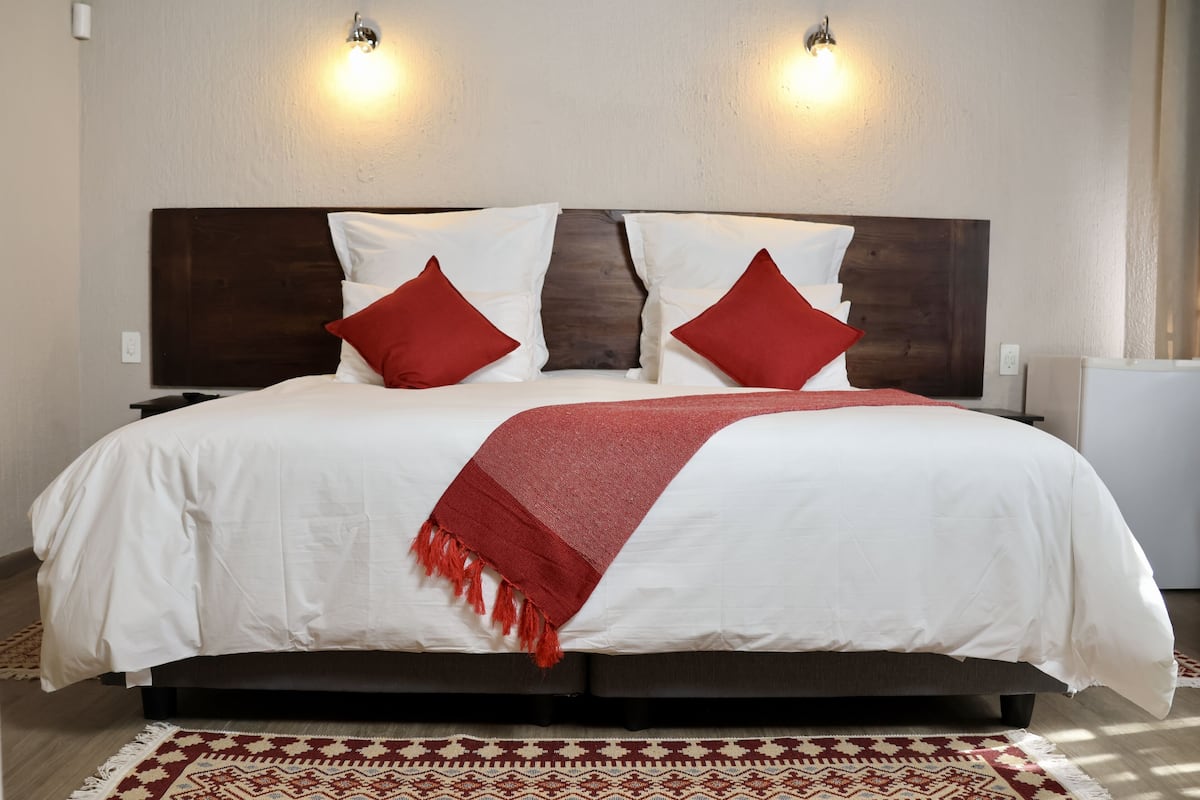 A neatly made bed with white bedding and red pillows, a red throw blanket, and a patterned rug on a wooden floor.