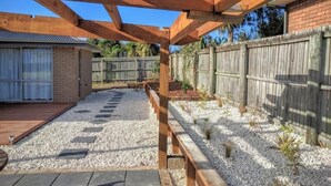 A backyard with a wooden deck, a brick house, a wooden fence, and a gravel path. - Spacious Quiet | 3BR | Decks | Wi-Fi | BBQ | Views (Kaikoura Township)
