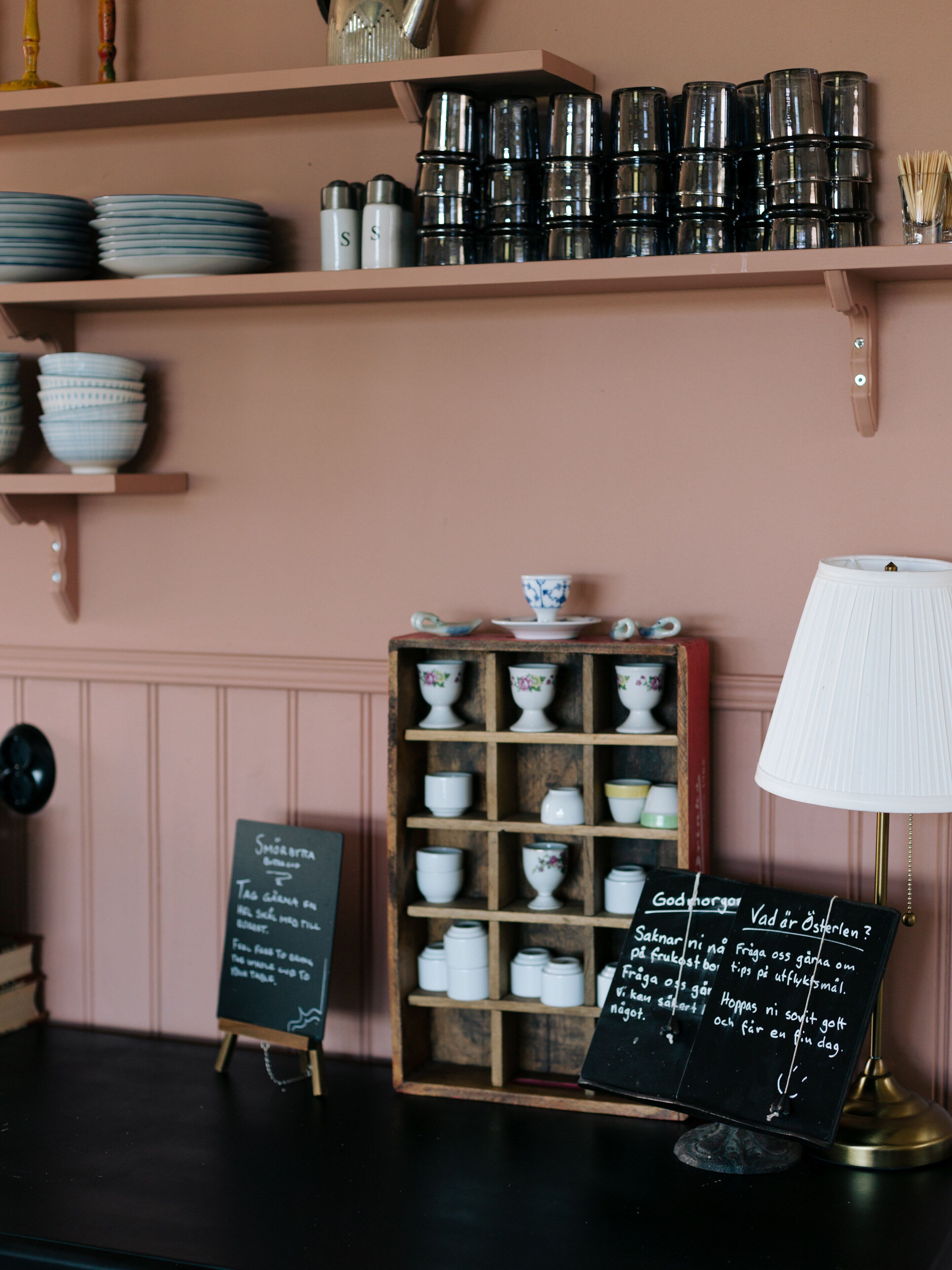A kitchen counter with shelves holding plates and jars, a wooden cabinet with bowls, and a chalkboard with writing.