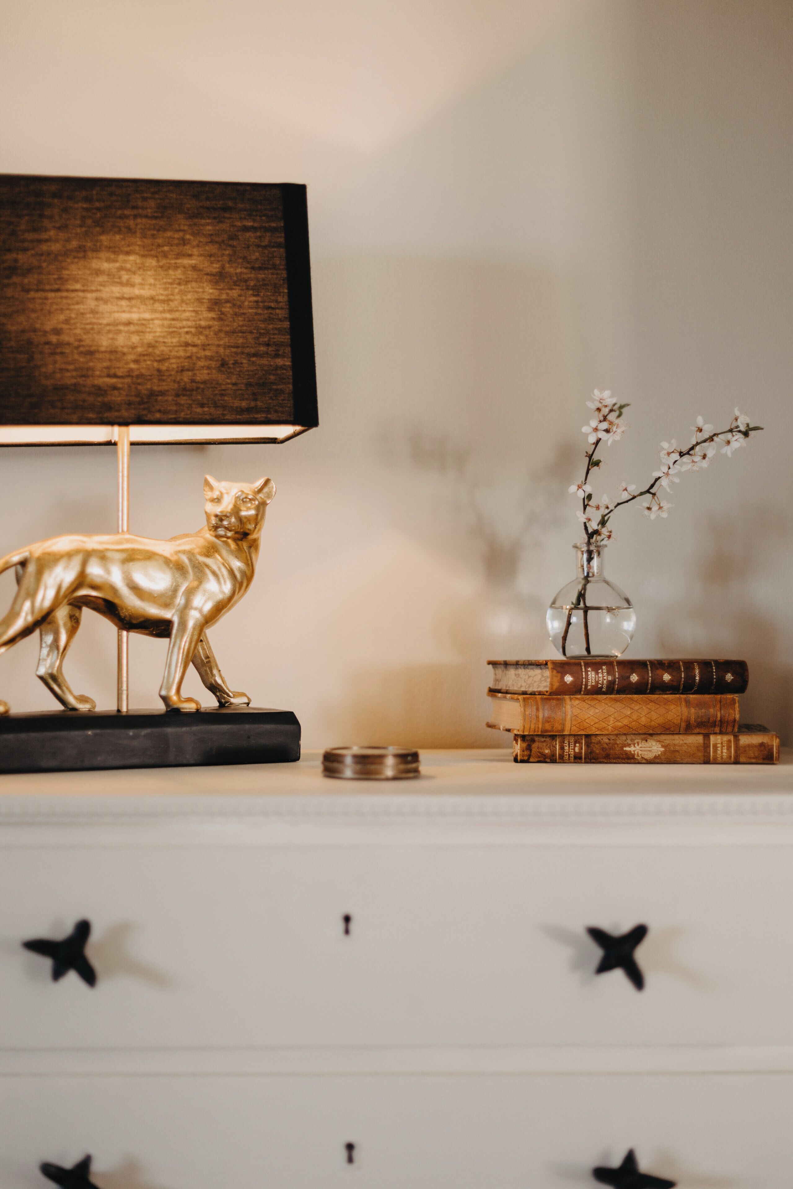 A white dresser with a gold cat figurine, a vase with flowers, and stacked books.