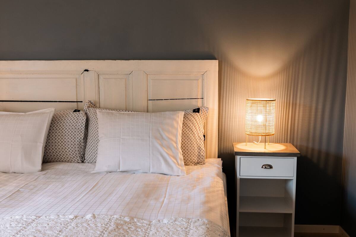A neatly made bed with a white comforter and pillows, a bedside table with a lamp, and a wooden floor.