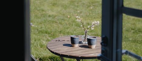 A wooden table with three cups and a vase with flowers, set on a deck with a vineyard in the background.