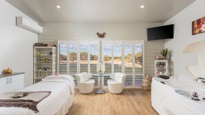 Um quarto de spa com uma cama, duas cadeiras brancas, uma mesa de massagem, uma banheira de hidromassagem e vista para o exterior.