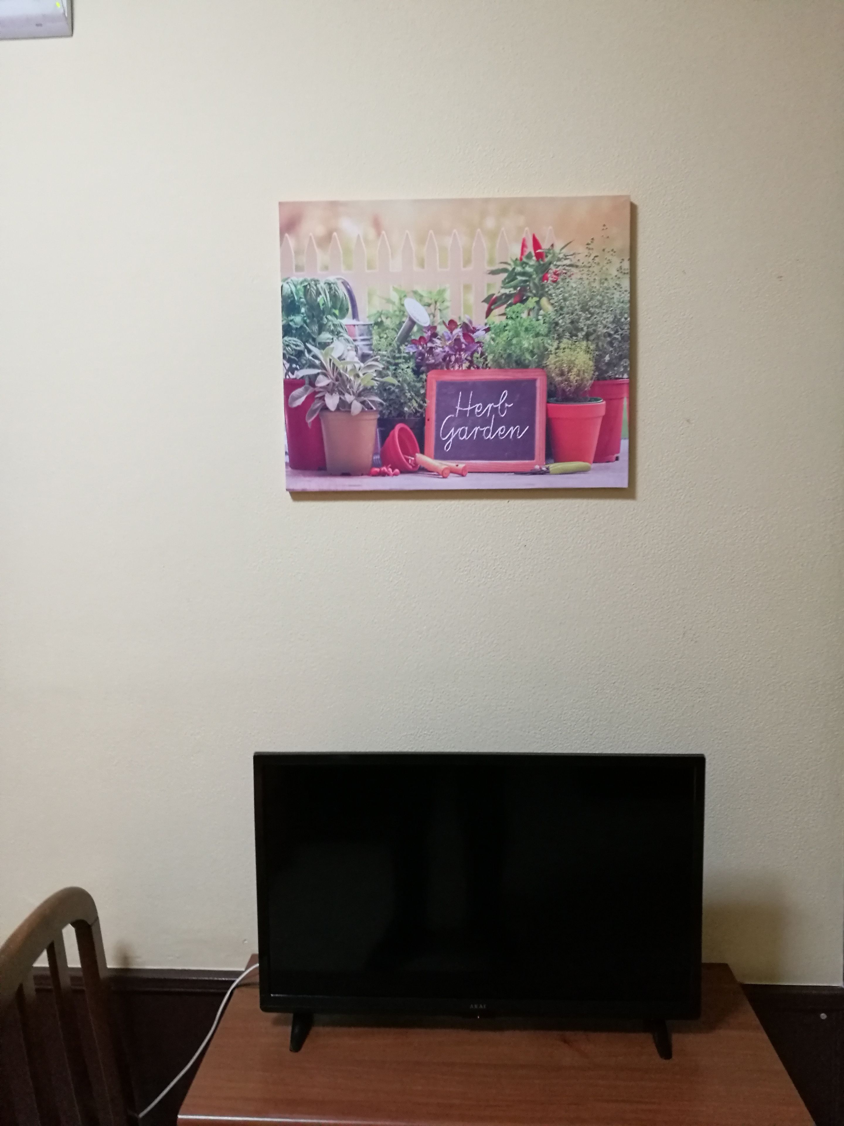 Een flatscreen televisie op een houten tafel, met een kamerplant en een bordje met de tekst 'Herb Garden' aan de muur achter de tafel.