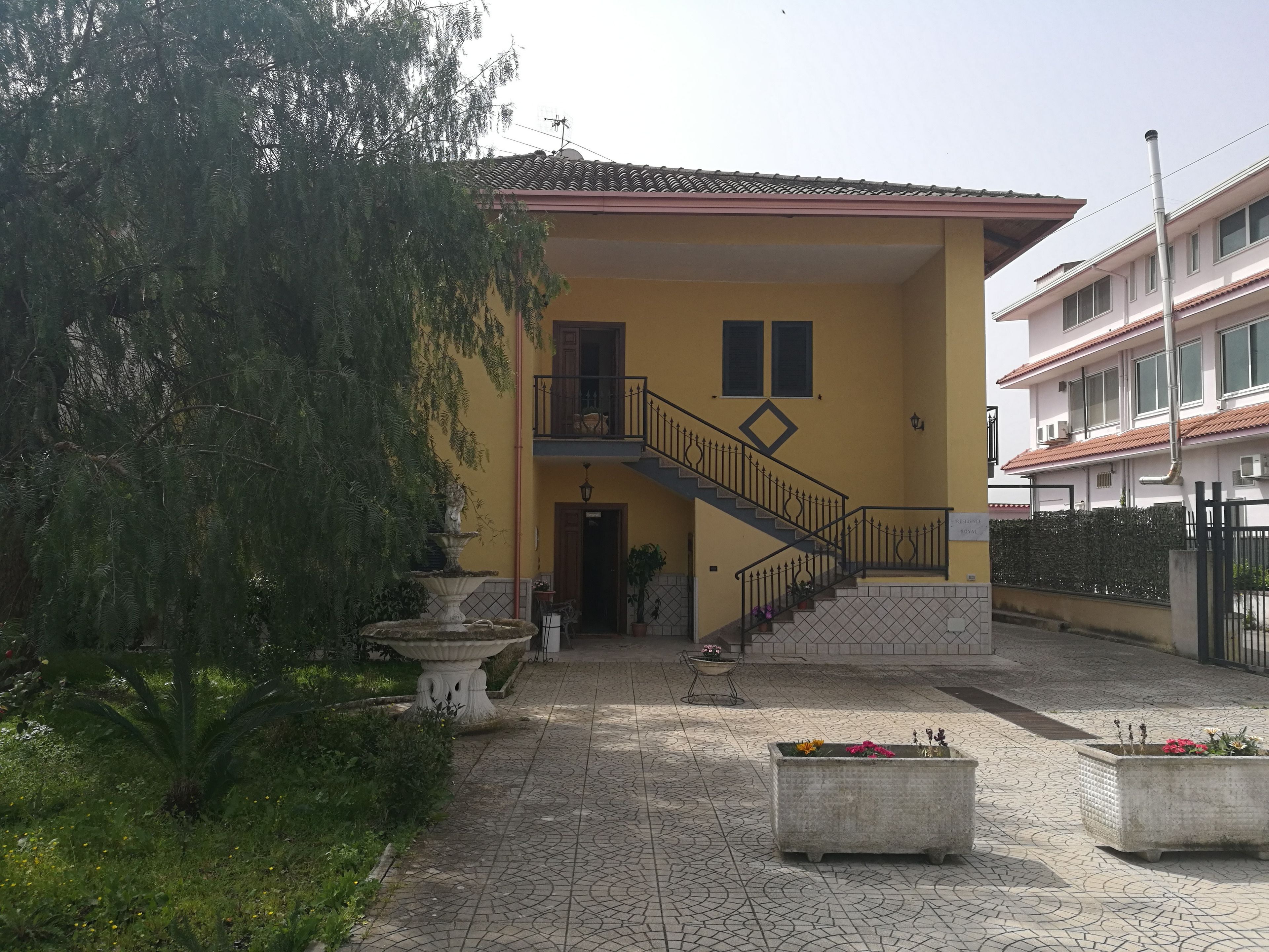 A yellow building with a staircase leading to a balcony, surrounded by greenery and potted plants.