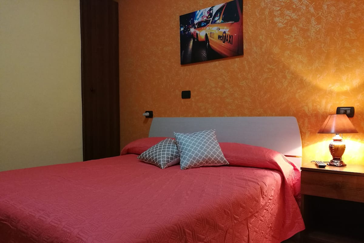 A hotel room with a bed, pillows, a nightstand with a lamp, and a wall-mounted picture.