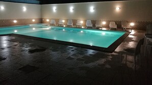 A swimming pool with clear water, surrounded by white lounge chairs and illuminated by overhead lights.