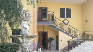 A yellow building with a stone fountain, a staircase with black railings, and potted plants.