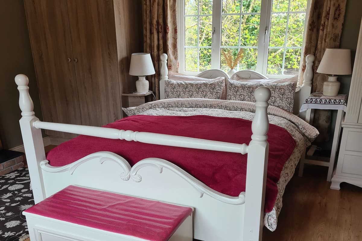 A bedroom with two beds, a window with curtains, a wooden wardrobe, and a bedside table with a lamp.