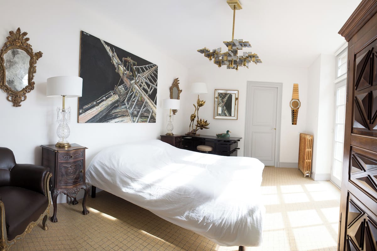 Ein Schlafzimmer mit Bett, Stuhl, Nachttisch, Gemälde, Spiegel, Lampe und Heizkörper.
