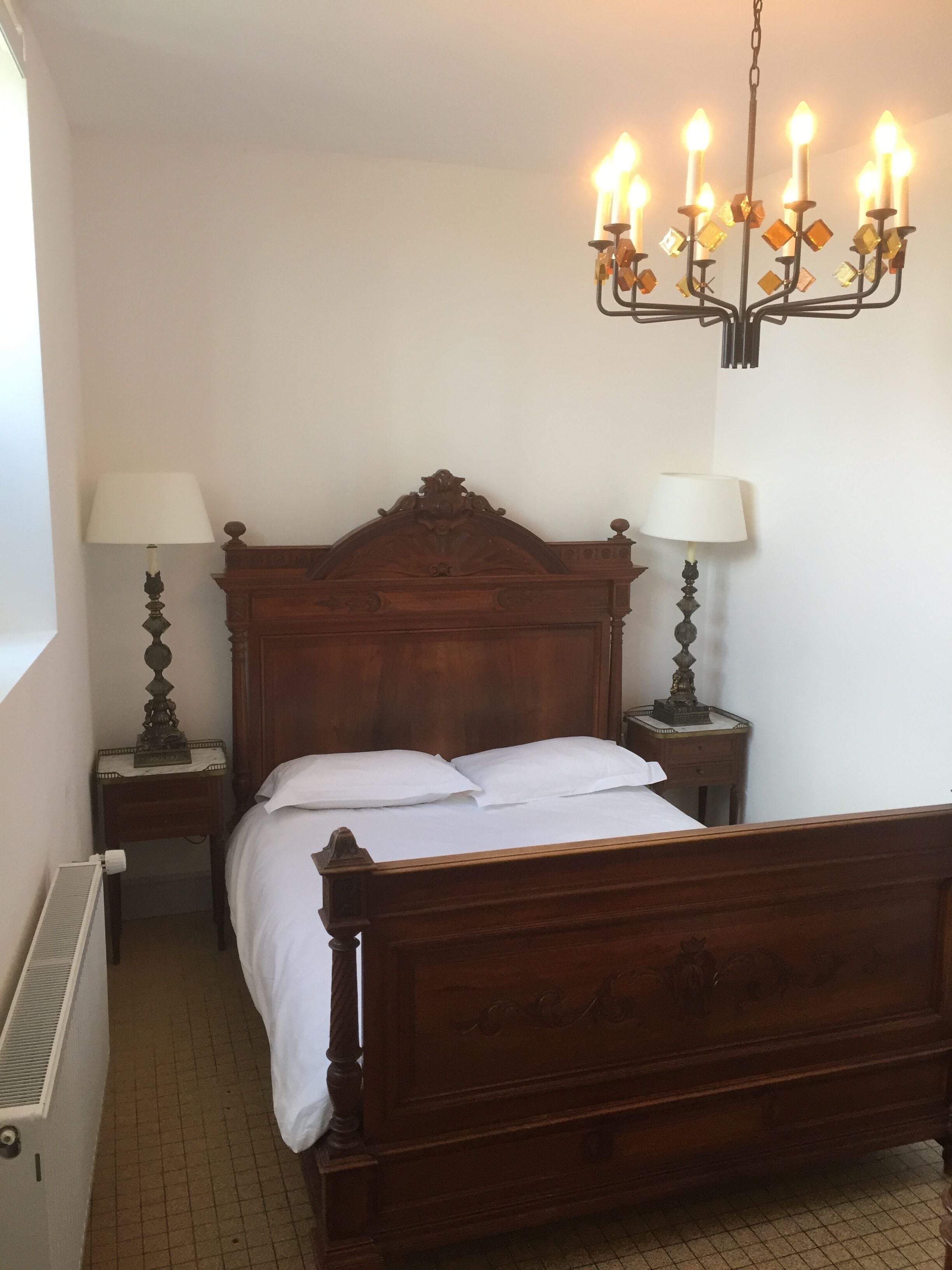 A bedroom with a wooden four-poster bed, two bedside tables with lamps, a radiator, and a chandelier.