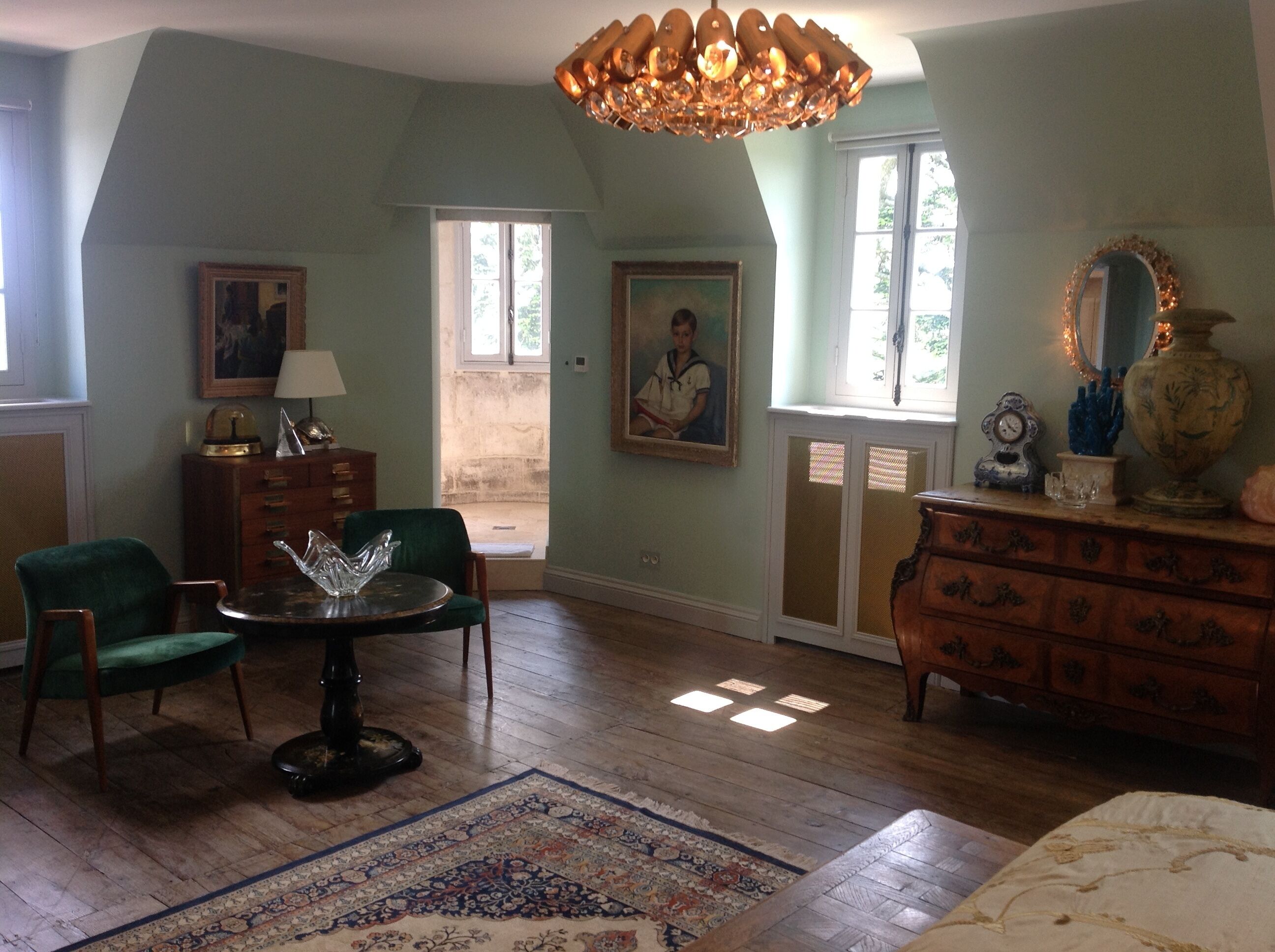 A room with a wooden floor, a green armchair, a table with a glass object, a dresser, and a chandelier.