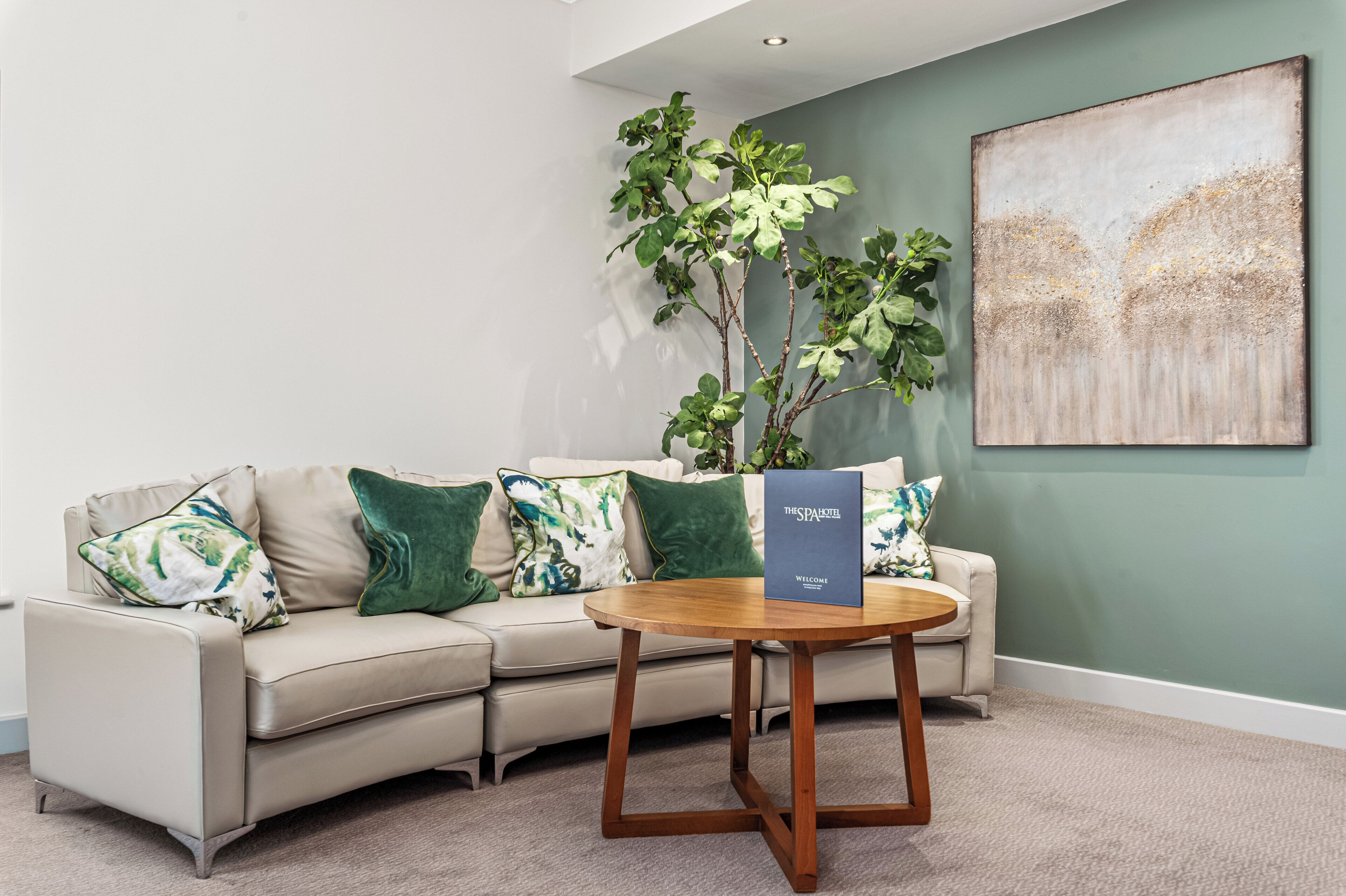 Uma sala de estar moderna com um sofá bege, almofadas estampadas em verde e branco, uma mesa de centro de madeira e um grande quadro abstrato na parede.