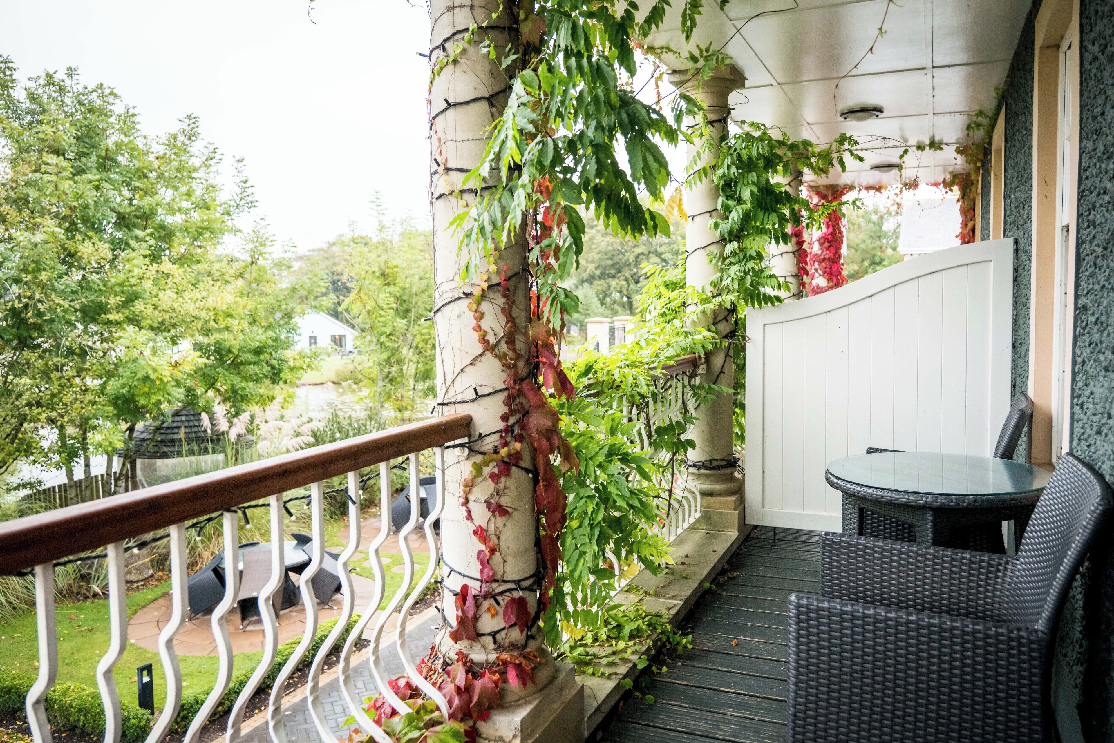Un balcon avec un fauteuil en osier, une balustrade blanche et une vue sur un jardin et une maison.