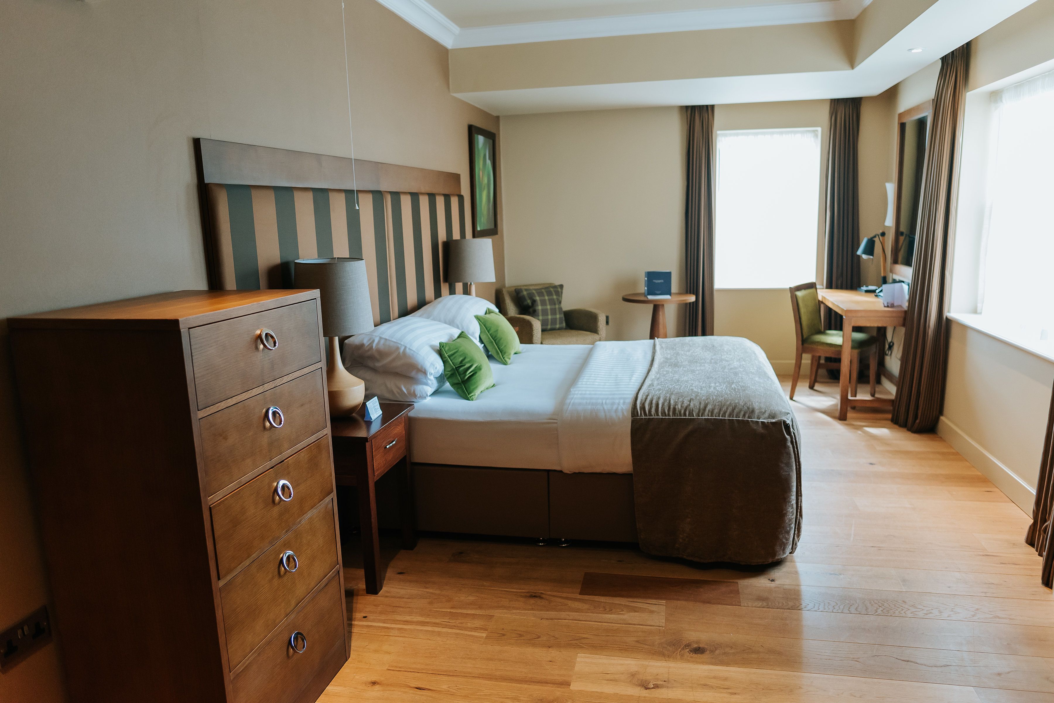 A hotel room with a bed, dresser, desk, chair, lamp, and curtains.