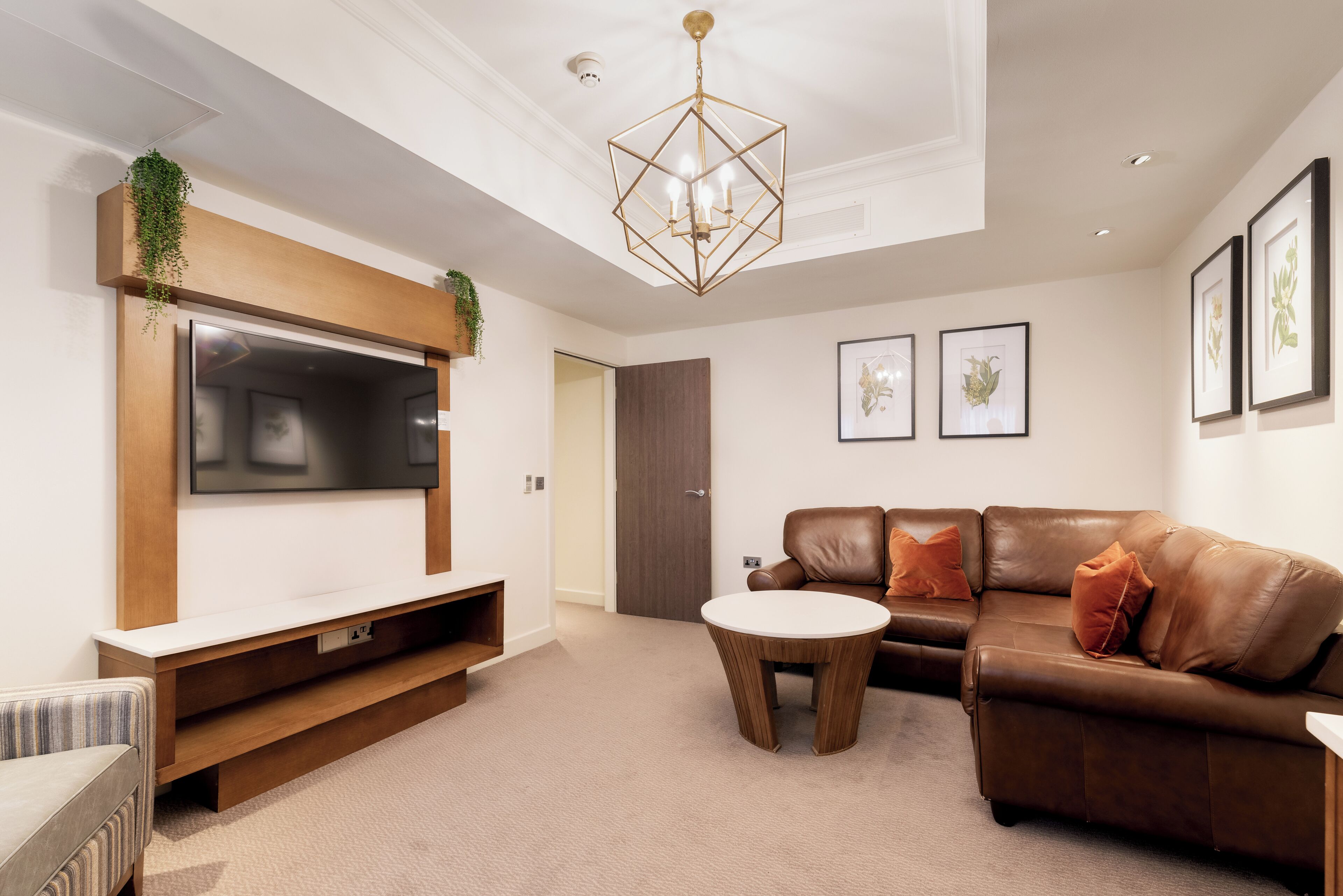 A modern living room with a flat-screen TV, a brown leather sofa, a round coffee table, and framed artwork on the walls.