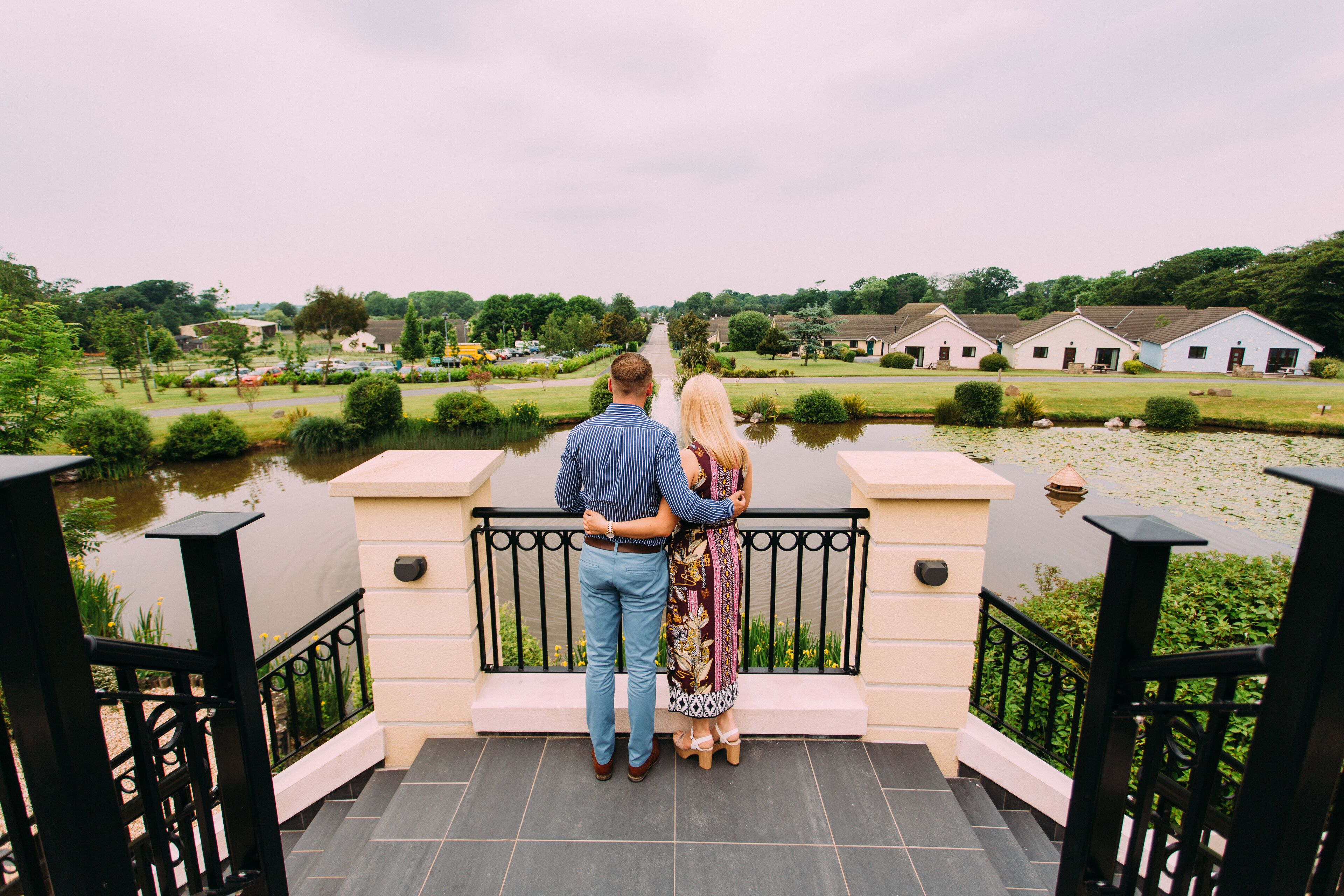A couple standing on a balcony overlooking a body of water, with a view of houses and greenery in the background.