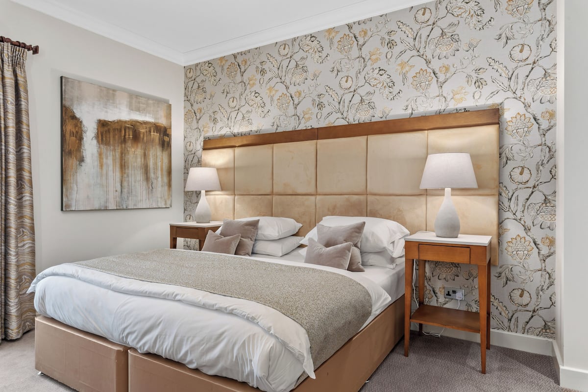 A neatly made bed with a headboard, two bedside tables with lamps, and a floral wallpaper pattern.
