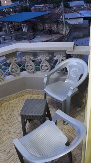 Un balcón con dos sillas de plástico y un taburete pequeño. El balcón tiene barandillas metálicas decorativas y vistas a edificios al fondo.