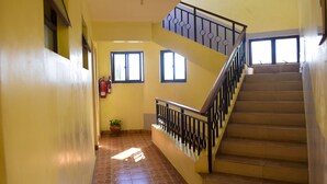 Un escalier avec une rampe métallique mène à l’étage supérieur. Des fenêtres sont disposées le long des murs, et on peut apercevoir un pot de fleurs sur la petite plateforme de l’étage inférieur.