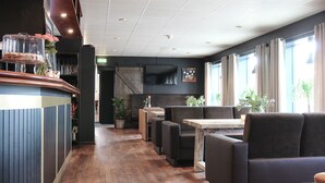 A restaurant with wooden flooring, black upholstered seating, a bar area with a counter, and large windows with curtains.